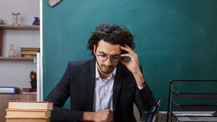 thinking-with-lowered-head-male-teacher-wearing-glasses-sitting-table-with-school-tools-classroom